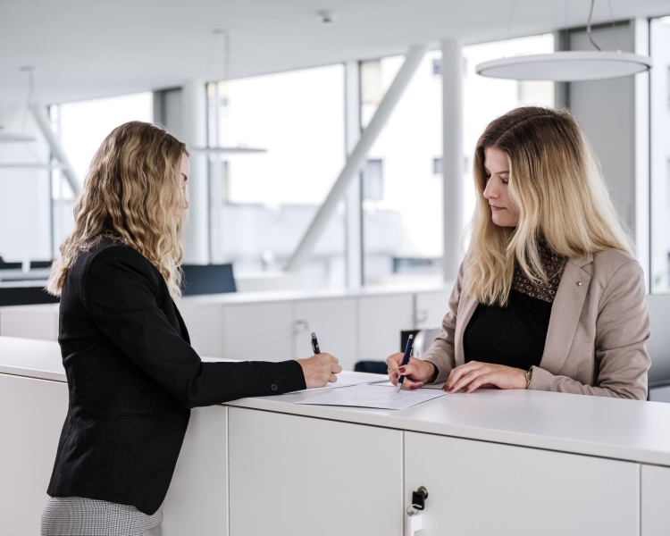 Zwei Frauen schreiben mit Kugelschreibern auf Dokumente auf einem Stehtisch