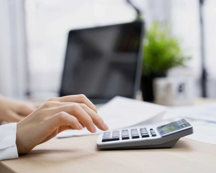 Eine Frau tippt auf einem Taschenrechner und im Hintergrund ist ein Computer zu sehen