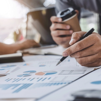 Ein Mann hält einen Kugelschreiber und betrachtet Unterlagen, eine Person im Hintergrund bedient ein Notebook