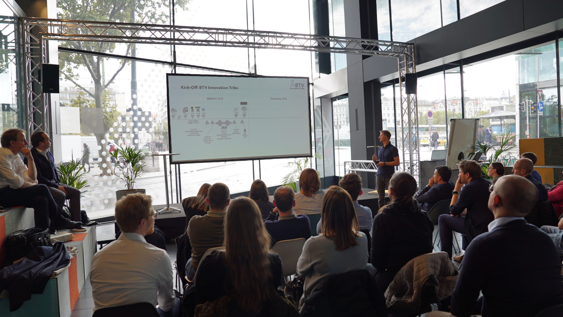 Ein Mann präsentiert auf einer großen Leinwand Power Point Folien, mehrere Zuschauer verfolgen diese Präsentation