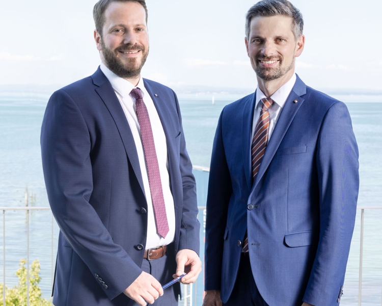 2 Männer lächeln in die Kamera und tragen jeweils eine blauen Anzug