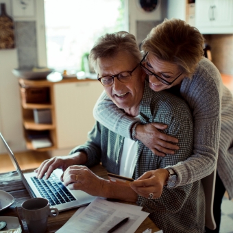 Eine Frau umarmt einen Mann der mit seinem Laptop an einem Tisch arbeitet