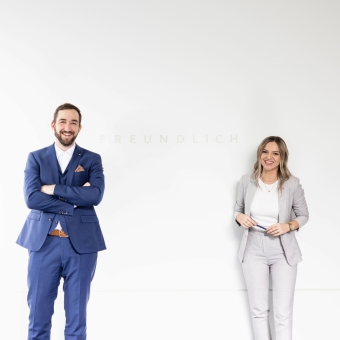 2 Personen stehen vor einer weißen Wand und tragen jeweils einen Anzug