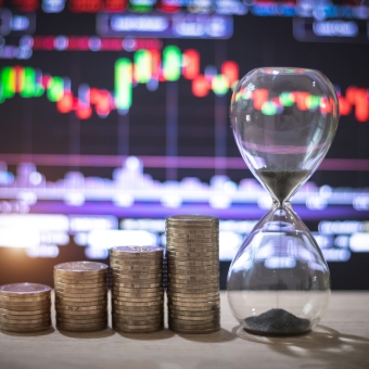 Vier Stapel Münzen stehen neben einer Sanduhr, dahinter ist ein Chart zu sehen, der die Zinsentwicklung zeigt