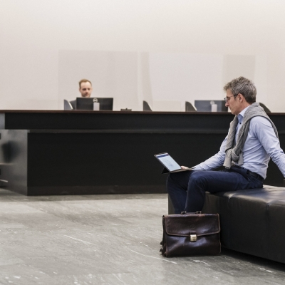 Ein Mann mit Brille sitzt auf einem Sessel in einer Bankfiliale und schaut auf seinem Tablet.