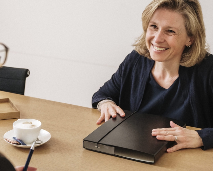 Eine Frau lächelt eine andere Frau an. Sie hat eine schwarze Mappe vor sich liegen.