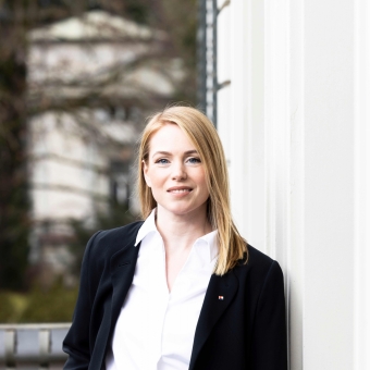 Eine Frau mit weißer Bluse und dunklem Blazer steht neben einer Gebäudewand, im Hintergrund befinden sich Bäume und ein weiteres Gebäude