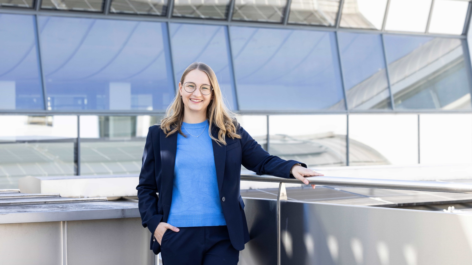 Eine Frau mit blonden Haaren, hellblauem Pullover und dunkelblauem Hosenanzug lächelt in die Kamera