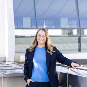 Eine Frau mit blonden Haaren, hellblauem Pullover und dunkelblauem Hosenanzug lächelt in die Kamera
