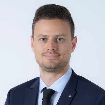 Ein Mann mit braunen Haaren trägt einen dunkelblauen Anzug, ein hellblaues Hemd und eine schwarzer Krawatte