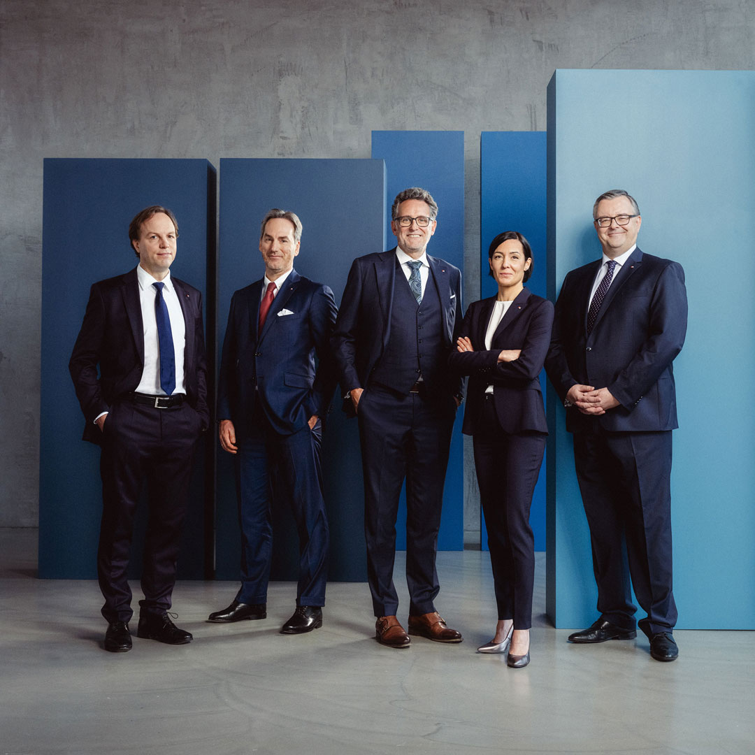 4 Männer und 1 Frau tragen einen Anzug mit einer blauen Wand im Hintergrund.