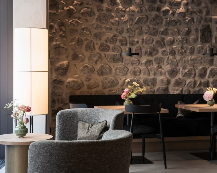 Tische und Stühle stehen in einem gut beleuchteten Raum mit Steinwand im Hintergrund. Auf den Tischen befinden sich Vasen mit Blumen.