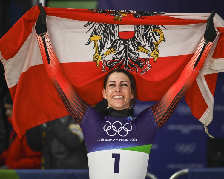 Skeletonpilotin Janine Flock hält eine Österreichflagge inkl. Tiroler Adler über ihren Kopf. Sie feiert ihre Goldmedaille auf den Olympischen Spielen in Milano Cortina 2026.