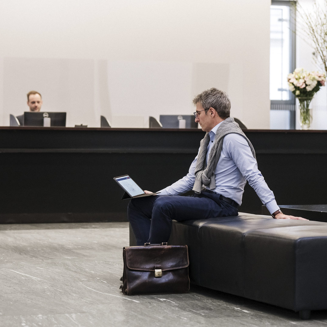 Ein Mann mit Brille sitzt auf einem Sessel in einer Bankfiliale und schaut auf seinem Tablet.