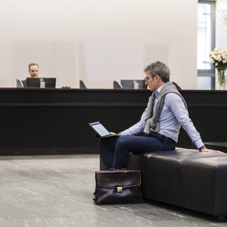 Ein Mann mit Brille sitzt auf einem Sessel in einer Bankfiliale und schaut auf seinem Tablet.