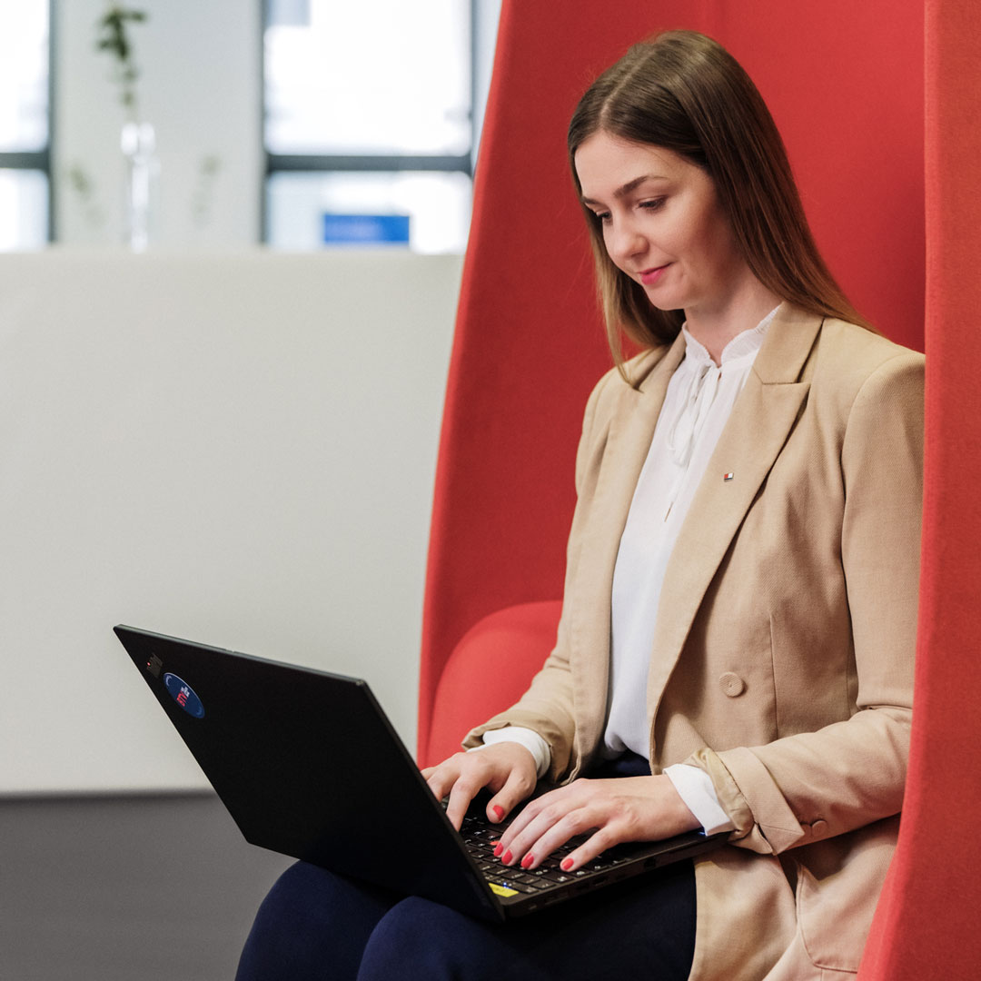 Eine Frau sitzt in einem roten Sessel und und arbeitet mit ihrem Laptop.